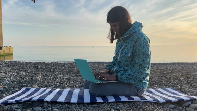 Work from anywhere. Digital nomad. Side view of a young woman, a freelance woman in cool weather, working at a laptop, sitting on the seashore. High quality 4k footage