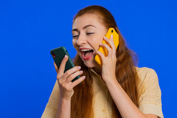 Irritated nervous stressed young woman talking screaming on two mobile phones having conversation conflict quarrel complaint dispute discuss solve problem. Girl isolated on blue studio background