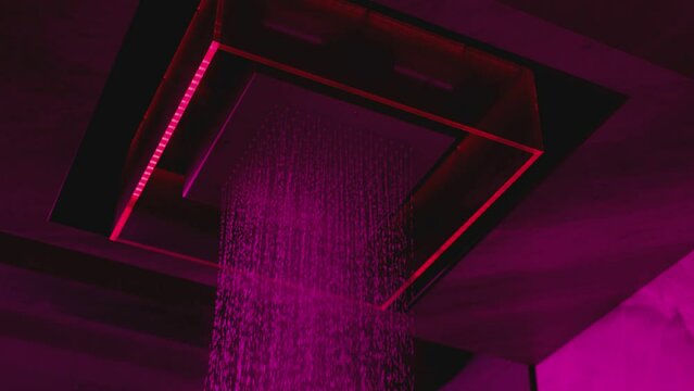 Water flowing from the beautiful modern designed shower . Overhead futuristic , luxury shower in dark bathroom . Pink , purple led lights inside . Unique design . Water falling , dropping , pouring