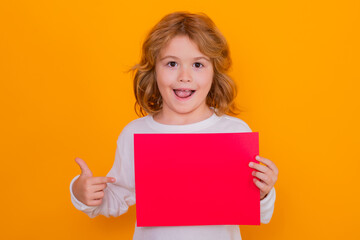 Fototapeta na wymiar Excited child boy showing index finger on red sheet of paper, isolated on yellow background. Portrait of a kid holding a blank placard, poster. Surprised face, amazed emotions of child.