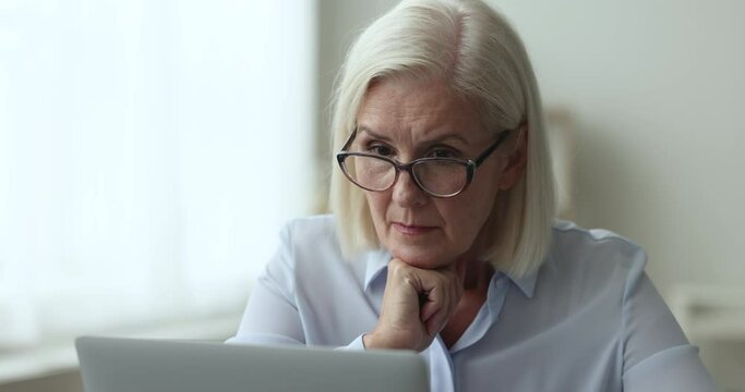 Focused senior businesswoman in elegant glasses working at laptop, typing, thinking on difficult decision, solving problem, looking at display, chatting with customer, leaning chin on hand