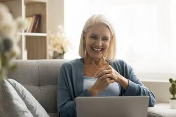 Happy blonde senior woman enjoying online connection on laptop at home, looking at computer,...