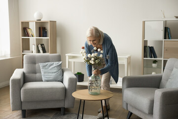 Happy blonde retired mature woman decorating home living room interior, placing flowers in glass...