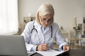 Serious senior head doctor woman working at office workplace, reading medical record, paper...