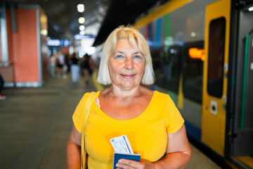Happy 50s senior woman biometric passport and train ticket waiting train on station platform....