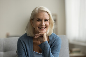 Cheerful pretty blonde senior woman looking at camera, smiling with healthy white teeth, laughing, posing for shooting on sofa, touching chin. Senior lady home head shot portrait - obrazy, fototapety, plakaty