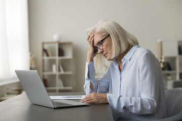 Tired mature senior business woman touching head at laptop, sitting at work table with computer,...
