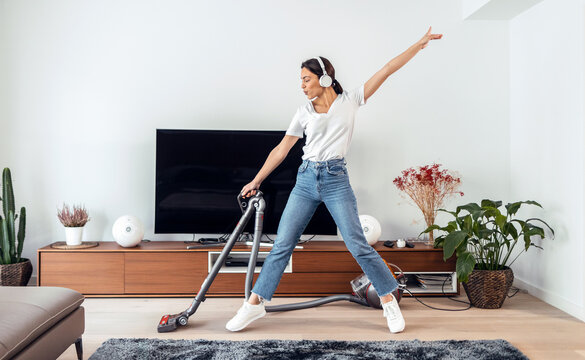 Young happy woman listening and dancing to music while cleaning the living room floor with a vaccum cleaner