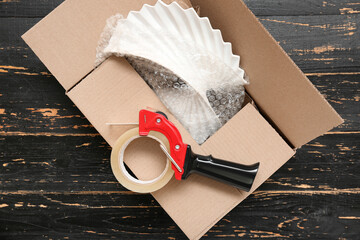 Packing tape dispenser and cardboard box on black wooden background
