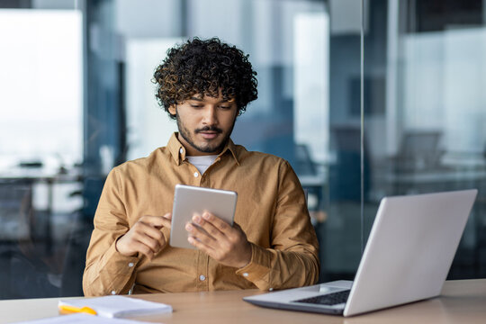 Serious focused businessman working with tablet computer sitting at desk inside office, hispanic thinking with laptop, man solving technical problem, using online application.