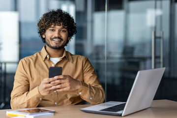 Portrait of a young successful Arab businessman inside the office, the man is smiling and looking at the camera, the employee is using an application on the phone, browsing online pages