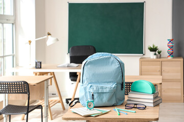 Blue school backpack with stationery, eyeglasses and alarm clock on desk in classroom