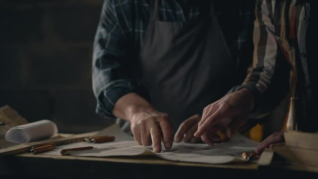 Handicraft Two colleagues workers studying blueprints of wooden parts for manufacturing At the workplace A young professional performs the task of making parts In a production room, small business