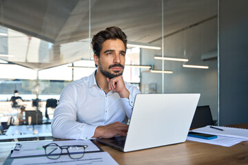 Fototapeta na wymiar Thoughtful young Latin business man looking away thinking using laptop at work. Serious busy professional businessman executive manager investor sitting in office planning investment strategy.