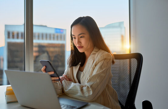 Young Busy Asian Business Woman Manager Using Laptop And Mobile Cell Phone Tech In Office. Professional Businesswoman Holding Smartphone, Working On Cellphone Looking At Computer Sitting At Desk.