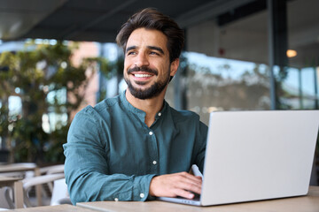 Smiling young Latin business man using laptop sitting outdoor. Happy guy student or professional looking away in city cafe elearning, hybrid working, searching job online thinking of digital solution. - obrazy, fototapety, plakaty