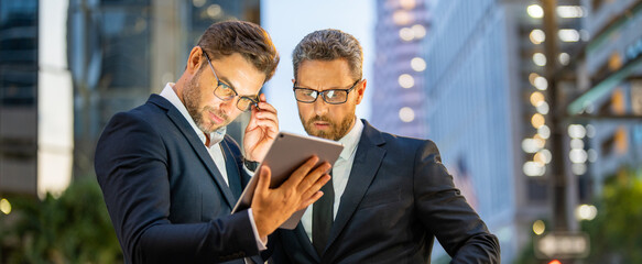 Discussing and planning remote working. Business men working on social media strategy with a digital tablet at night city. Two businessman outdoors sharing tablet, remote working, discussing.