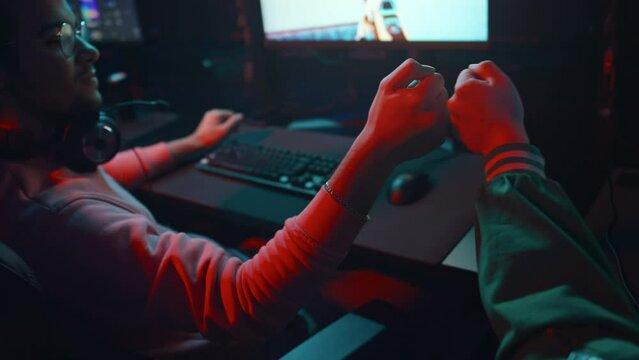 Close-up rear slowmo shot of two young male friends playing first person shooter game together in cyber cafe, winning and bumping fists