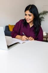 Vertical shot of young indian student woman taking notes while using laptop computer at home. Millennial ethnic female learning online listening virtual video call. Business and education concept.