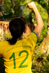 Torcedora brasileira de costas com a blusa verde amarela numero 12, comemorando o gol com soco no ar e ao fundo a natureza. © Madu Oliveira