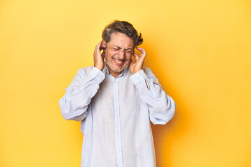 Middle-aged man posing on a yellow backdrop covering ears with hands.