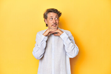 Middle-aged man posing on a yellow backdrop keeps hands under chin, is looking happily aside.