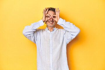 Middle-aged man posing on a yellow backdrop showing okay sign over eyes