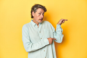 Middle-aged man posing on a yellow backdrop shocked pointing with index fingers to a copy space.