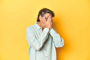 Middle-aged man posing on a yellow backdrop blink through fingers frightened and nervous.