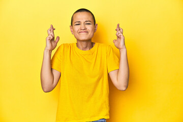 Young woman in yellow tee, yellow studio backdrop crossing fingers for having luck