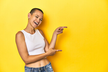 Shaved head woman in white tank top, yellow backdrop pointing with forefingers to a copy space,...