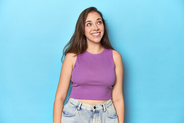 Fashionable young woman in a purple top on blue background relaxed and happy laughing, neck stretched showing teeth.