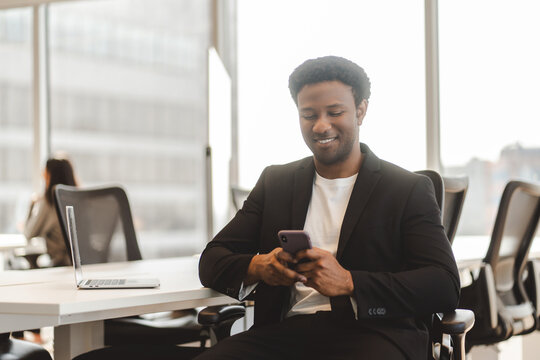 Attractive Middle Aged African American Man Holding Mobile Phone Reading Text Message In Modern Office. Pensive Guy Sitting With Smartphone Using Mobile App Shopping Online, Ordering Food