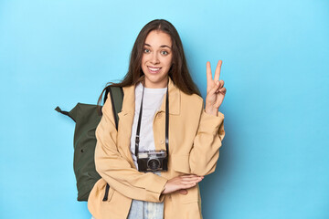 Young Caucasian woman traveler with camera and backpack showing number two with fingers.