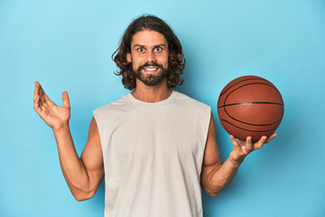 Bearded man with basketball in blue studio surprised and shocked.