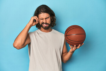 Bearded man with basketball in blue studio covering ears with hands.