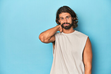 Bearded man in a tank top, blue backdrop touching back of head, thinking and making a choice.