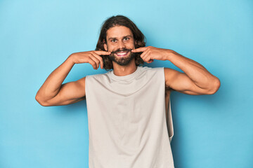 Bearded man in a tank top, blue backdrop smiles, pointing fingers at mouth.