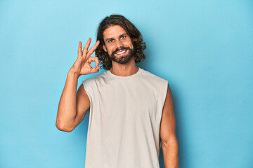 Bearded man in a tank top, blue backdrop cheerful and confident showing ok gesture.