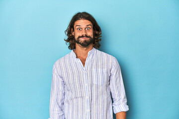 Fototapeta premium Man with beard in blue striped shirt, blue studio blows cheeks, has tired expression. Facial expression concept.