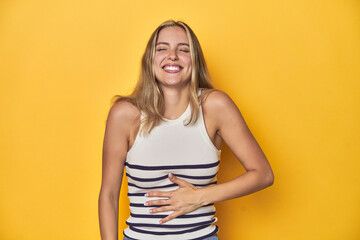 Obraz premium Young blonde Caucasian woman in a white tank top on a yellow studio background, touches tummy, smiles gently, eating and satisfaction concept.