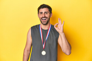 Athletic man with gold medal, yellow studio backdrop cheerful and confident showing ok gesture.