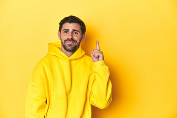 Man in yellow hoodie, monochrome studio backdrop showing number one with finger.