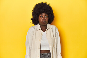 African-American woman with afro, studio yellow background shrugs shoulders and open eyes confused.