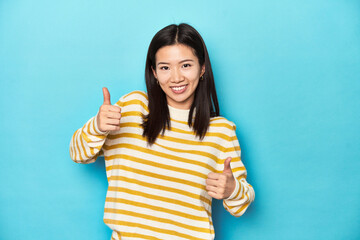 Asian woman in striped yellow sweater, raising both thumbs up, smiling and confident.