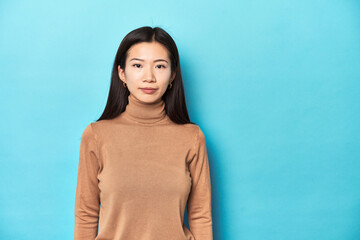 Asian woman in brown turtleneck looking at camera, blue background.