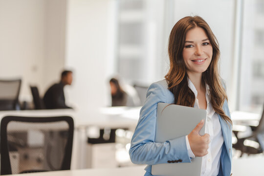 Smiling confident woman wearing stylish business suit holding laptop, looking at camera at modern office. Good looking businesswoman in formal wear posing for picture. Concept of successful business 