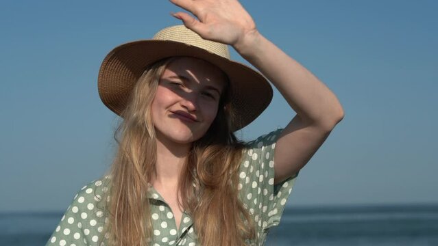 Authentic young woman in straw hat and light green polka dot dress on background of sea waves on sunny summer day. 21 year old European female enjoying sunshine and breeze. Cinematic shot, slow motion