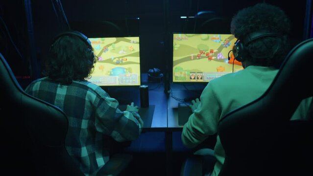 Medium rear shot of unrecognizable young girl and guy sitting in cyberclub, playing fun anime cartoon style, real time strategy video game together, scoring and bumping fists