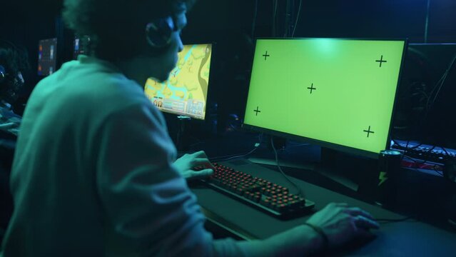 Medium rear shot of unrecognizable young man with curly hairdo playing video game on computer with green chromakey screen, girl playing cartoon game in background. Suitable as template, copy space